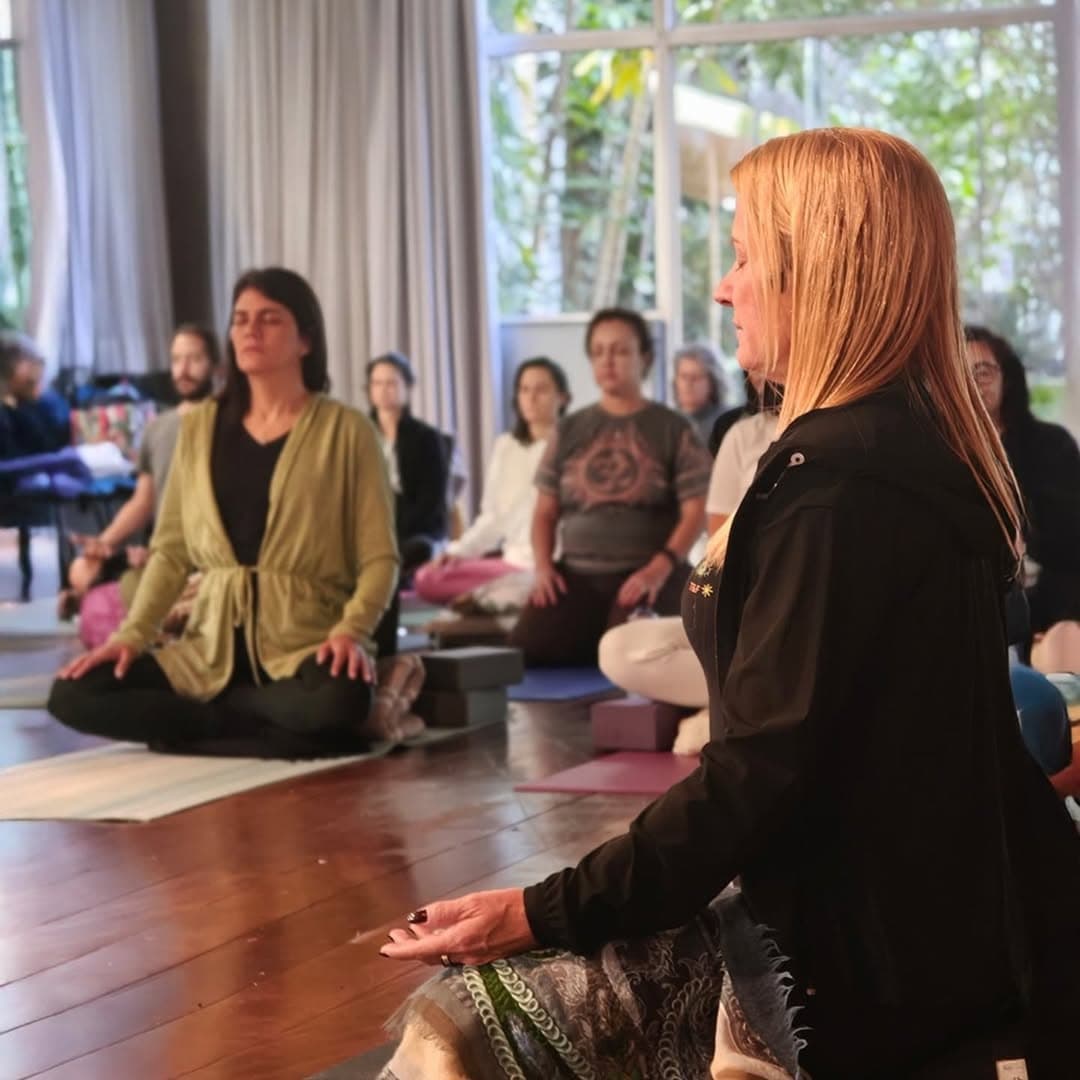 Deni Miranda conduzindo uma sala em meditação, com alunas sentadas em lótus